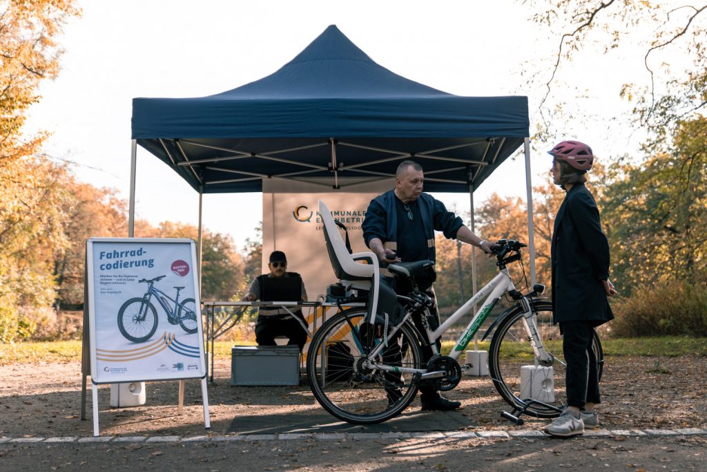 Ein Mann und eine Frau stehen an einem Fahrrad und sind im Gespräch. Im Hintergrund ein blauer Pavillon und ein Schild "Fahrradcodierung Leipzig"