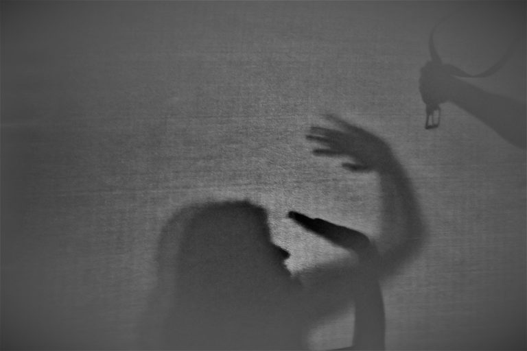 Silhouette. Eine Frau schützt ihr Gesicht mit den Händen. Von oben kommt eine Hand mit einem Gürtel ins Bild.