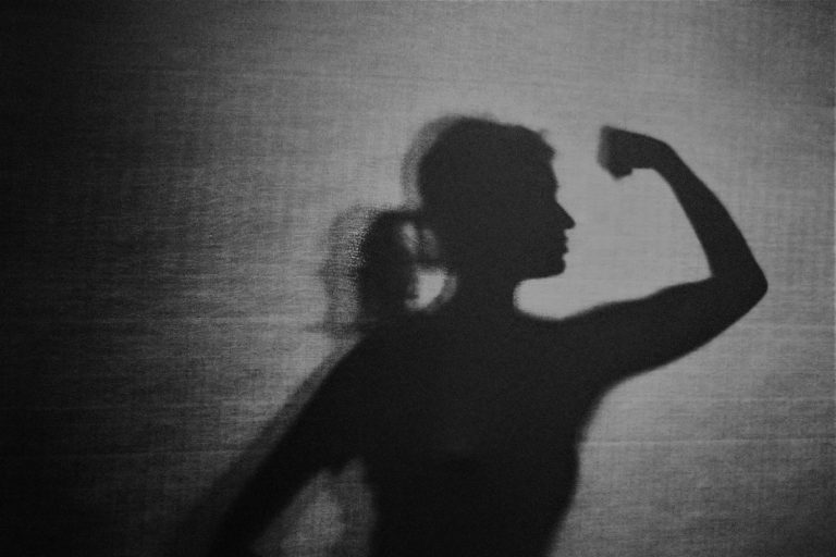Silhouette: Eine Frau steht in starker Pose, einen Arm nach vorne gestreckt, am Ellbogen 90 Grad angewinkelt, die Hand zur Faust geballt.