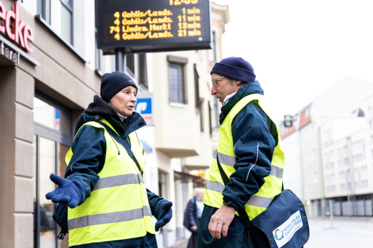 Zwei Personen in gelben Warnwesten unterhalten sich. Im Hintergrund die Anzeigetafel einer Straßenbahnhaltestelle.
