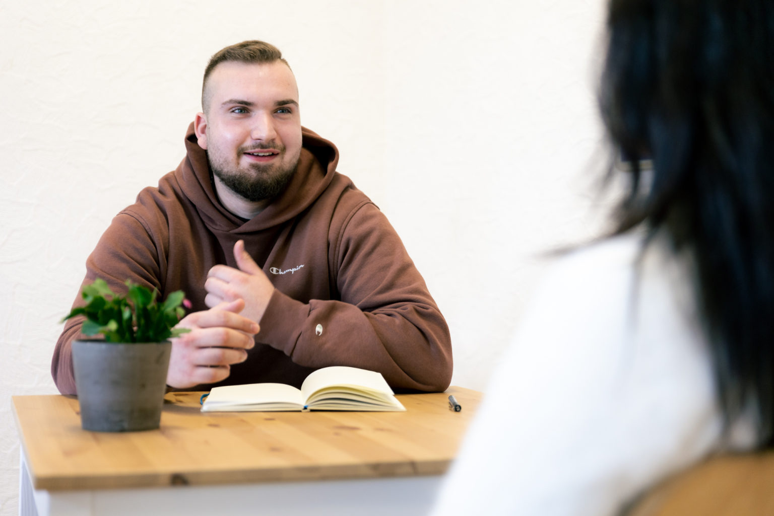 Ein junger Mann sitzt an einem Tisch einer nicht erkennbaren Person gegenüber und redet mit ihr.
