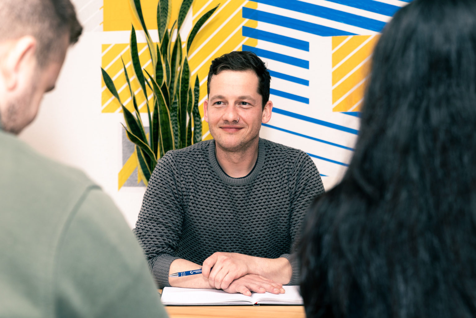 Ein Berater sitzt zwei nicht erkennbaren Personen gegenüber. Sie reden. Er hat einen Stift in der Hand und ein Notizbuch vor sich liegen.