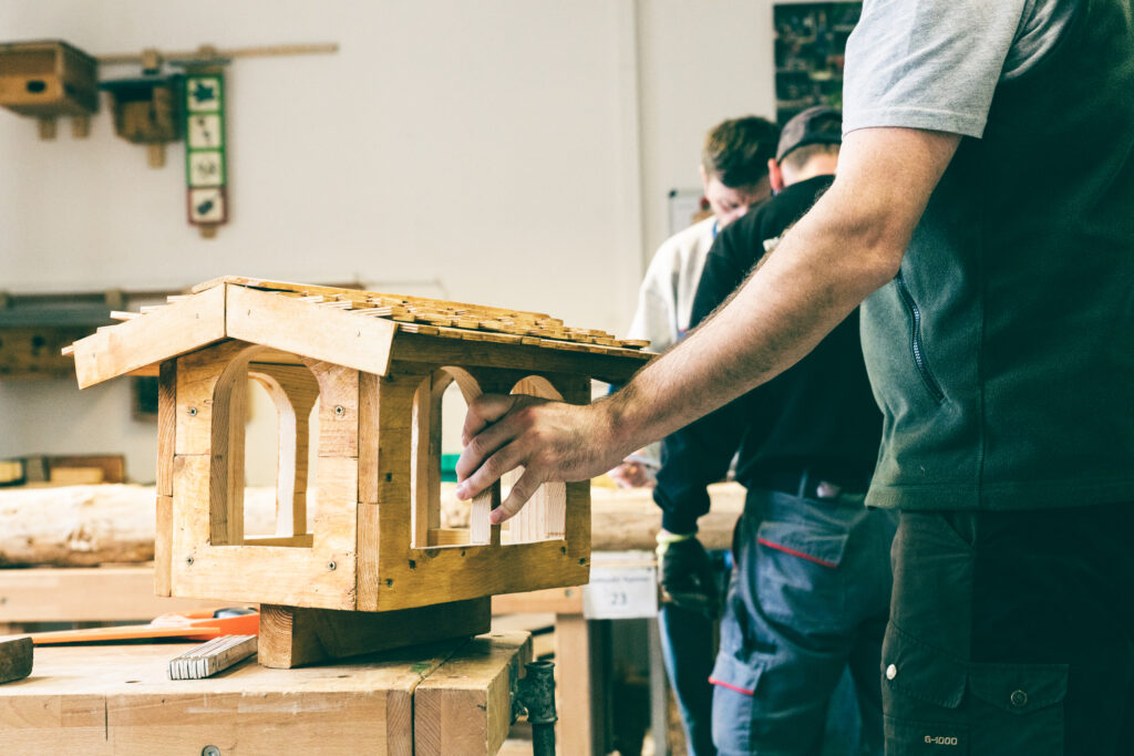 Ein Hand hält ein Vogelhaus fest, dass auf einer Werkbank in einer Holzwerkstatt steht.