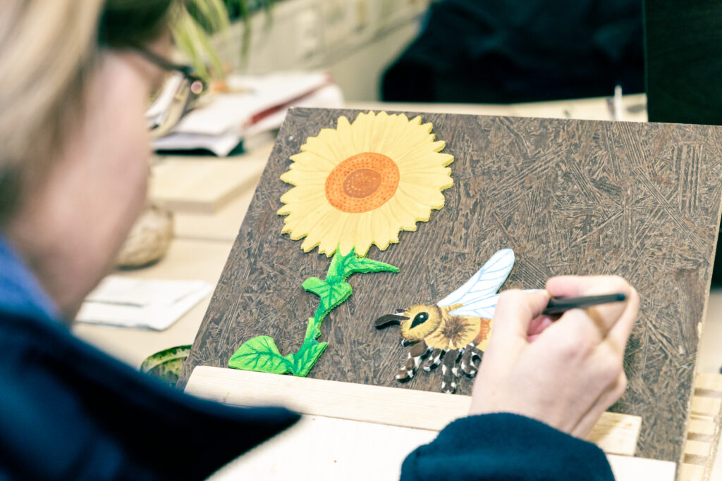 Eine Person, verschwommen im Vordergrund, bemalt mit einem Pinsel eine Sonnenblume und eine Biene aus Holz.