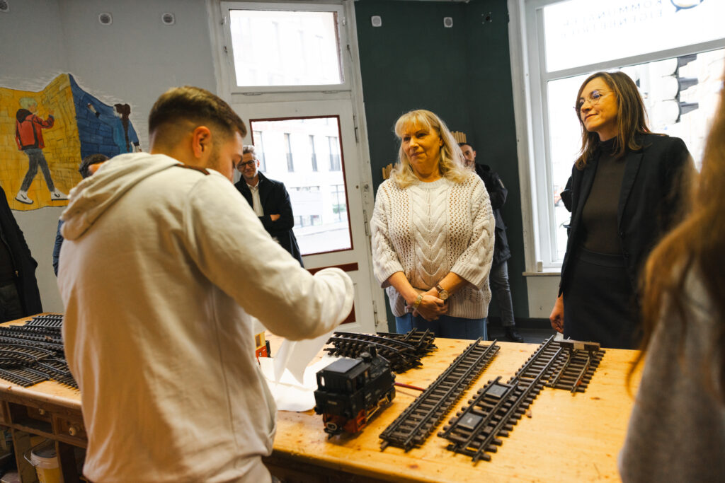 Ein Projektteilnehmer erläutert Staatsministerin Petra Köpping und Sozialbürgermeisterin Vicki Felthaus Arbeiten an einer Modelleisenbahn für Außenbereiche, die in der S.A.V.E. Projektwerkstatt instandgesetzt wird.