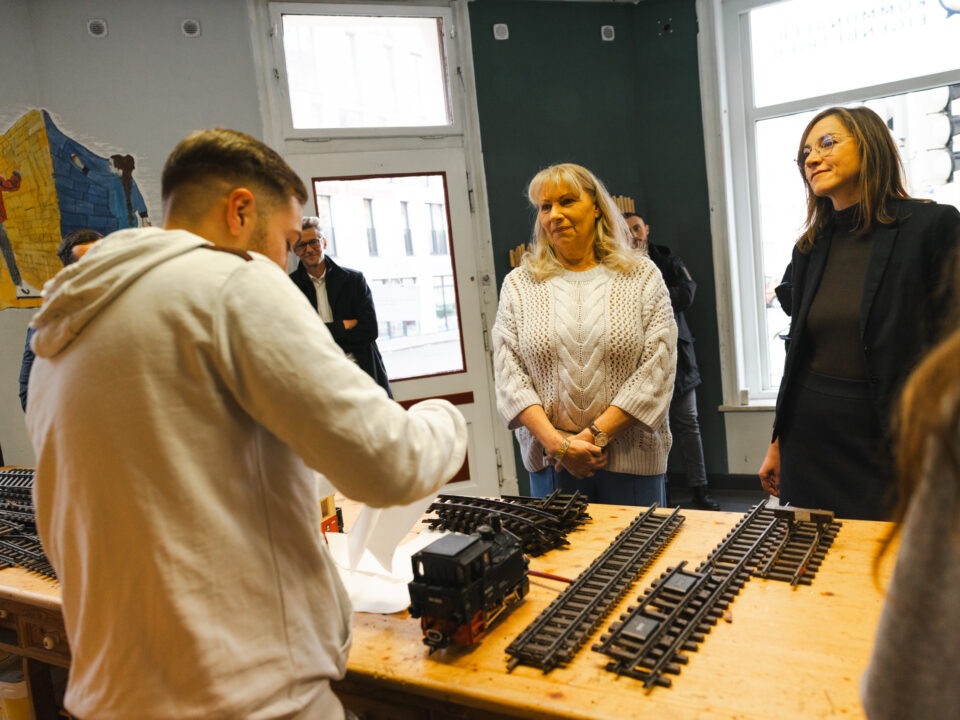 Ein Projektteilnehmer erläutert Staatsministerin Petra Köpping und Sozialbürgermeisterin Vicki Felthaus Arbeiten an einer Modelleisenbahn für Außenbereiche, die in der S.A.V.E. Projektwerkstatt instandgesetzt wird.